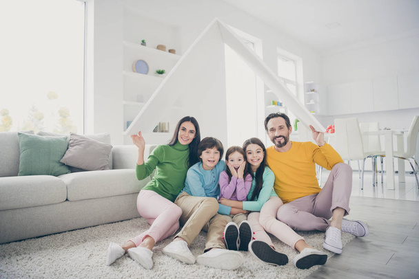 Portrait of nice attractive lovely excited cheerful family three pre-teen kids sitting on carpet floor holding in hand roof place good change at light white interior style house flat living-room - 写真・画像