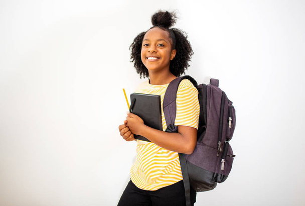 Портрет африканской девочки-подростка, улыбающейся с сумкой и книгами на изолированном белом фоне
 - Фото, изображение