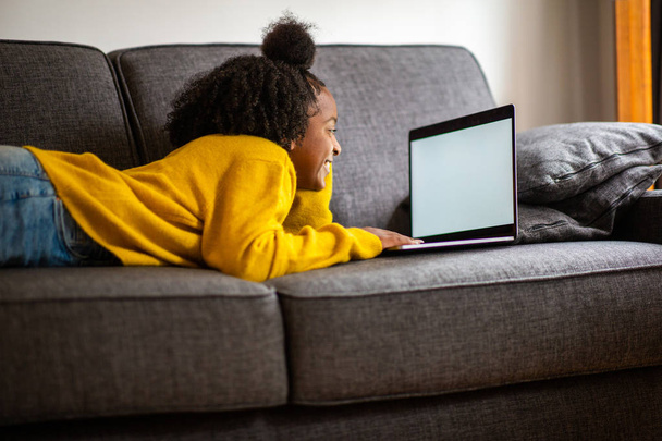 Portrait african american girl lying on sofa looking at laptop computer screen at home - 写真・画像