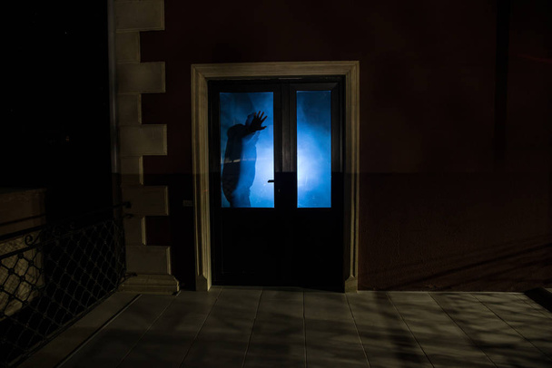 Silhouet van een onbekende schaduwfiguur op een deur door een gesloten glazen deur. Het silhouet van een mens voor een raam 's nachts. Enge scène halloween concept van wazig silhouet van maniak. - Foto, afbeelding