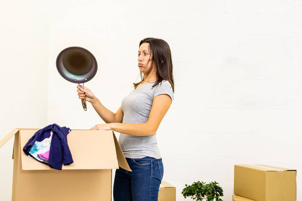 Brunette caucasian woman unpacking boxes, holding a frying pan, looking at it in confusion. - Fotoğraf, Görsel