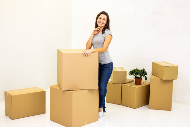Happy woman prepared all carton boxes for moving into new flat and stands leaning on a pile of boxes, smiling to the camera. - 写真・画像