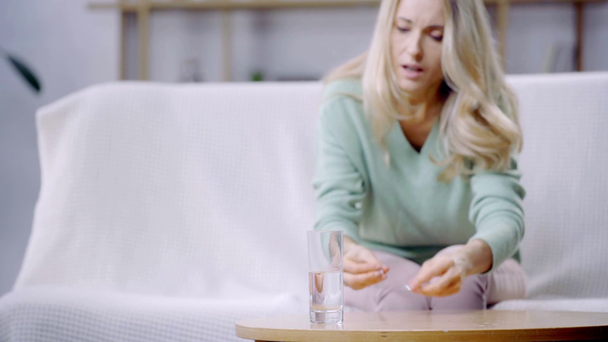 επιλεκτική εστίαση του γυαλιού του νερού και της γυναίκας με πονοκέφαλο λαμβάνοντας χάπι στο παρασκήνιο  - Πλάνα, βίντεο