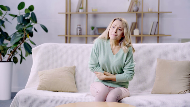 kobieta siedzi na kanapie i cierpi na ból brzucha - Materiał filmowy, wideo