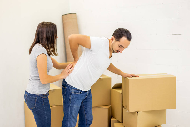 Man suffering from cramp in back while stacking boxes, preparing for relocation, woman tries to help him. - 写真・画像