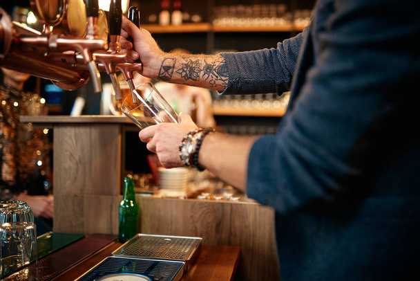 Tattooed caucasian barman pouring beer while standing in pub. Selective focus on hand. - 写真・画像