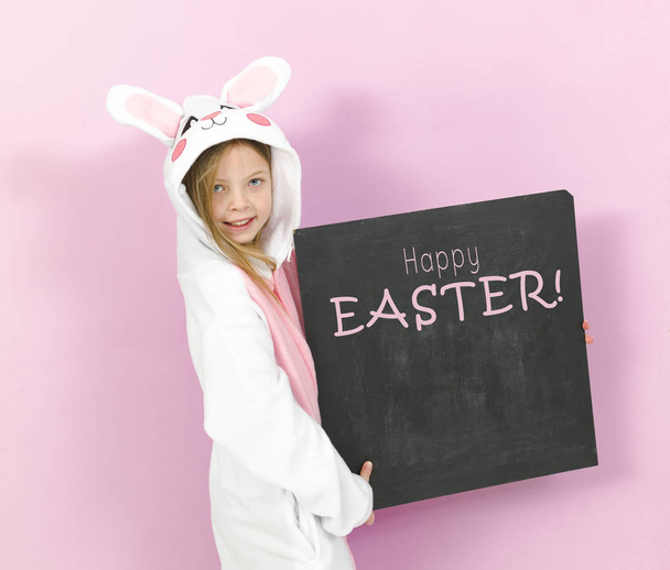 pretty blonde girl in cozy rabbit costume holding white board with text happy easter and posing in studio  - Valokuva, kuva