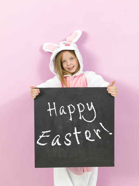 pretty blonde girl in cozy rabbit costume holding white board with text happy easter and posing in studio  - Fotografie, Obrázek