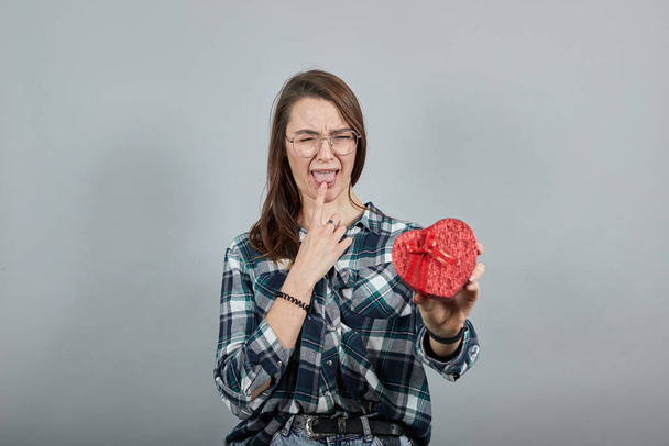 upset woman in glasses holds gift box in shape of heart, touches finger tongue - Fotoğraf, Görsel