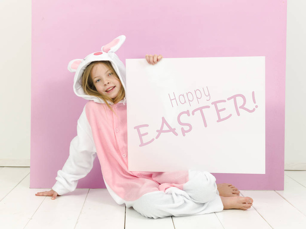 pretty blonde girl in cozy rabbit costume holding white board with text happy easter and posing in studio  - Valokuva, kuva