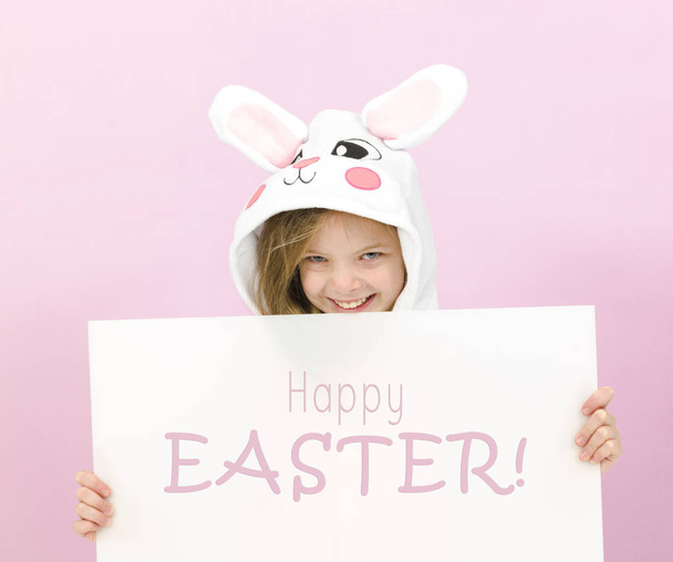 pretty blonde girl in cozy rabbit costume holding white board with text happy easter and posing in studio  - Foto, Imagem