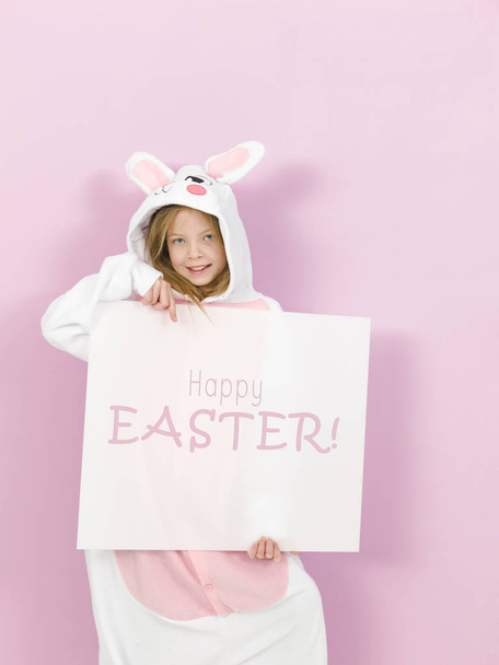 pretty blonde girl in cozy rabbit costume holding white board with text happy easter and posing in studio  - Fotografie, Obrázek