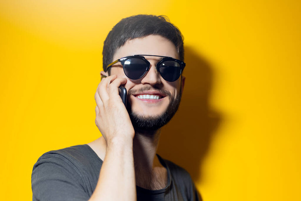 Studio portrait of young smiling man using smartphone, wearing sunglasses on yellow background. - 写真・画像