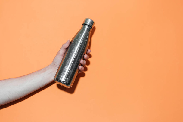 Close-up of male hand holding a steel reusable thermo eco bottle for any liquid, sprayed with water, isolated on orange background. - Valokuva, kuva
