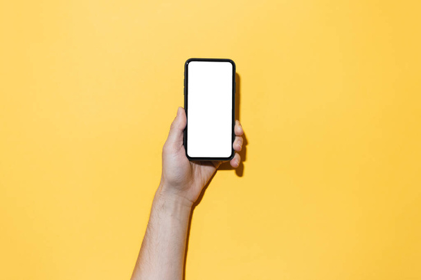 Крупный план мужчины, держащего в руках смартфон с пустым белым макетом, изолированный на желтом фоне
. - Фото, изображение