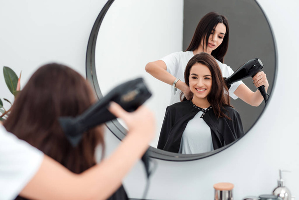 Милая красивая девушка с длинными волосами смотрит в зеркало, она пришла в салон красоты, ее парикмахер работает с ее волосами
. - Фото, изображение