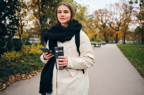 Pretty casual girl in down jacket walking around park with cellphone and thermo cup - Foto, Imagen