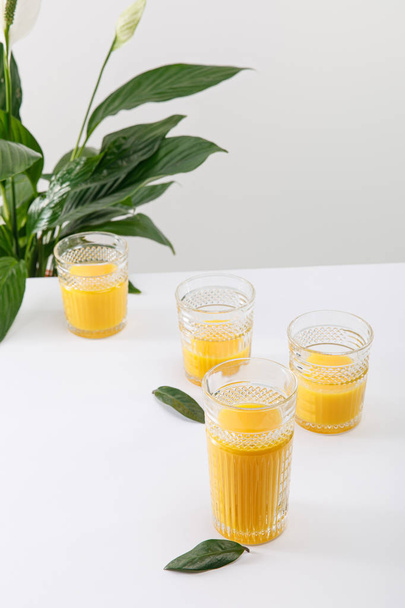 copos de smoothie amarelo delicioso fresco na superfície branca perto de planta de lírio paz verde isolado em cinza
 - Foto, Imagem