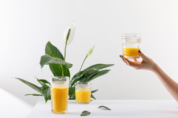 cropped view of woman holding glass of fresh delicious yellow smoothie near green peace lily plant on white - Фото, изображение
