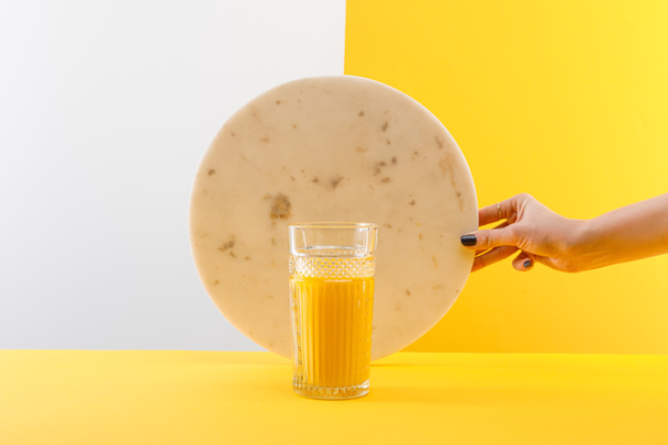 oříznutý pohled na ženu držící mramorový kruh v blízkosti skla čerstvého lahodného žlutého smoothie na šedém a žlutém pozadí - Fotografie, Obrázek