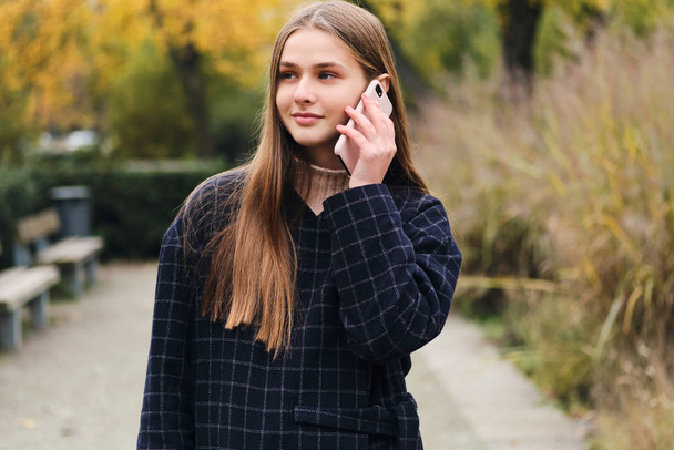 Pretty smiling girl in coat talking on cellphone walking alone in park - Фото, изображение