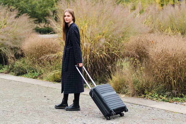 Beautiful stylish girl in coat with suitcase dreamily looking in camera outdoor - Foto, Imagem
