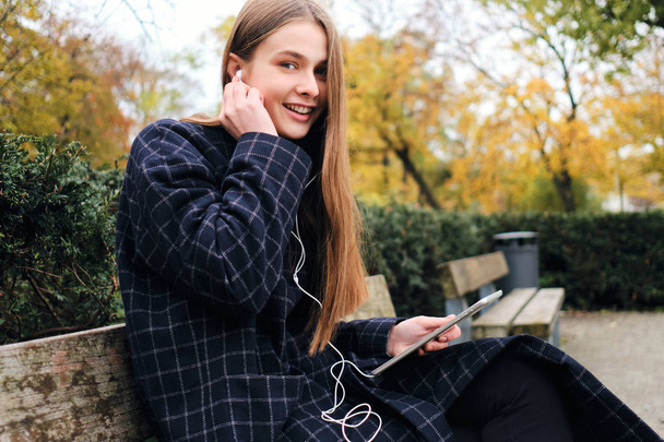 Pretty cheerful girl in earphones happily resting with tablet on bench in city park - Фото, изображение