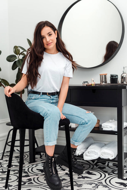 Charming girl in a white T-shirt and jeans sits on a chair near a hairdressing table, in a beauty salon - Фото, изображение