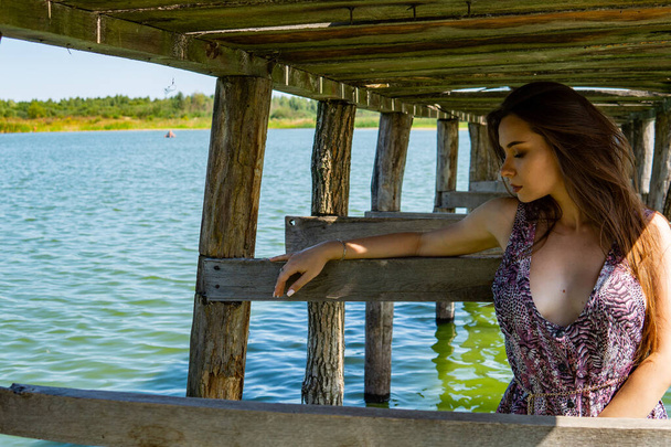Dark-haired woman posing at the wooden pier at the lake. Woman in summer outfit with her back turned to the wooden footbridge of Lake Neusiedl. Sun reflections at the boathouse in the background. - Photo, Image