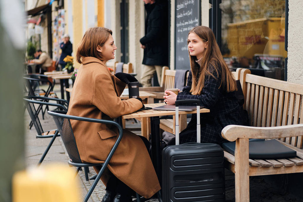2人の美しいスタイリッシュな女の子が夢のようにスーツケースと屋外カフェで一緒に休んで話をする - 写真・画像