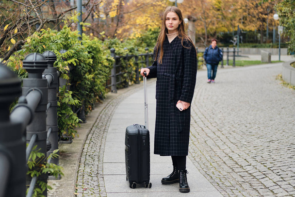 Beautiful casual girl in coat intently looking in camera with little black suitcase on street - Photo, Image