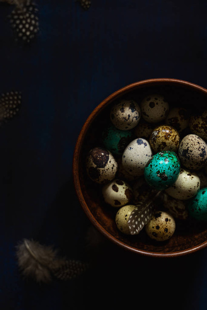Перепелиные яйца, цветной чирок, перья в миске на темно-синем фоне. Тёмная капризная пасхальная открытка
 - Фото, изображение