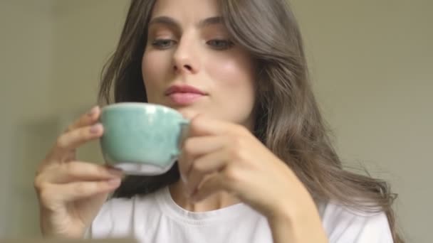A beautiful young woman is drinking coffee and looking to the window while using a laptop computer indoors - Materiał filmowy, wideo