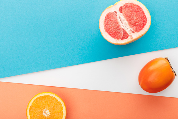 Horní pohled na grapefruitovou půlku, plátek citrusových plodů a persimmon na modrém, oranžovém a bílém pozadí - Fotografie, Obrázek