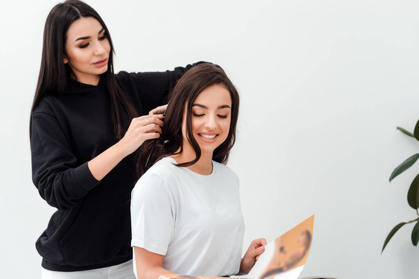 Smiling, attractive brunette is reading a magazine while her hairdresser combs her beautiful hair - Fotoğraf, Görsel