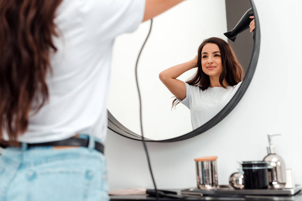 Όμορφη νεαρή γυναίκα στεγνώνει τα όμορφα μαλλιά της και κοιτιέται στον καθρέφτη - Φωτογραφία, εικόνα