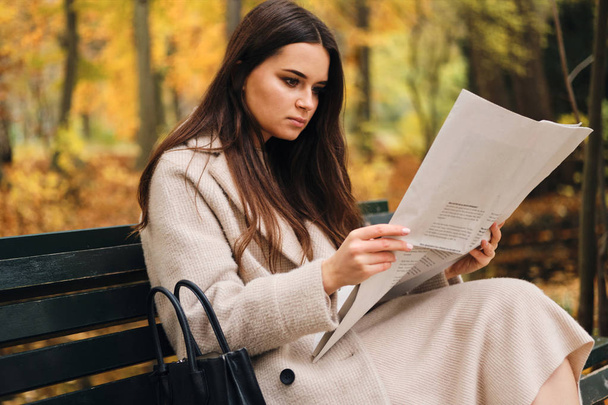 mooi brunette meisje aandachtig lezen krant zitten alleen op bank in de herfst park  - Foto, afbeelding