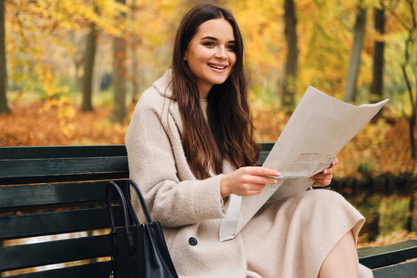 Pretty uśmiechnięta brunetka dziewczyna szczęśliwie czyta gazetę na ławce w jesiennym parku miejskim  - Zdjęcie, obraz