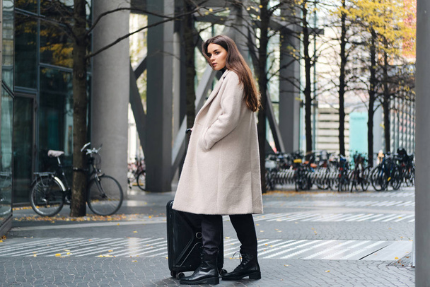 Ελκυστικό κομψό κορίτσι με αυτοπεποίθηση κοιτάζοντας στην άκρη στέκεται στο δρόμο της πόλης με μικρή βαλίτσα - Φωτογραφία, εικόνα