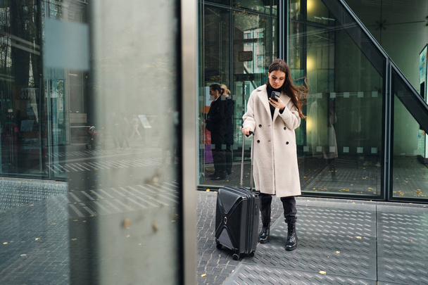 Νεαρή όμορφη επιχειρηματίας με παλτό χρησιμοποιώντας κινητό τηλέφωνο περιμένει στο δρόμο με βαλίτσα - Φωτογραφία, εικόνα
