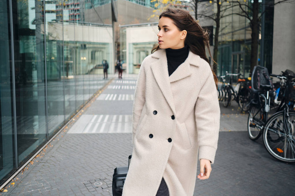 Young attractive businesswoman in coat confidently walking around city street with suitcase - Fotoğraf, Görsel