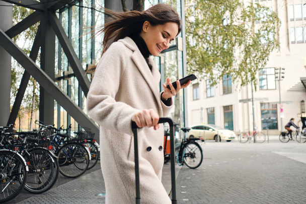 Mladý pěkný businesswoman v kabátě šťastně pomocí mobilního telefonu procházky ulicí s kufrem - Fotografie, Obrázek
