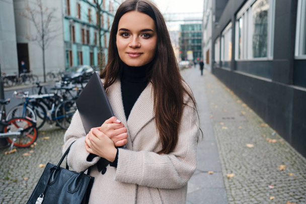 Beautiful smiling brunette girl with laptop happily looking in camera on city street - Valokuva, kuva