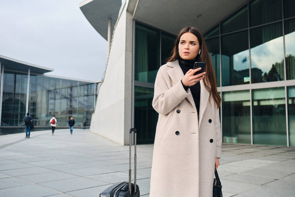Joven mujer de negocios con estilo en abrigo cuidadosamente usando el teléfono celular esperando en la calle de la ciudad con la maleta
 - Foto, imagen