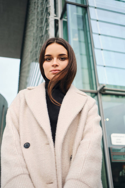 Portrait of beautiful brunette girl in coat confidently looking in camera in business district of city - Foto, Imagem