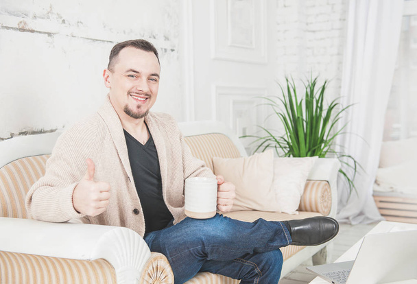 Bonito homem sorridente com xícara de café sentado no sofá em casa
 - Foto, Imagem
