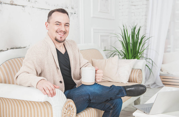 Jóképű mosolygós férfi egy csésze kávéval otthon ül a kanapén. - Fotó, kép
