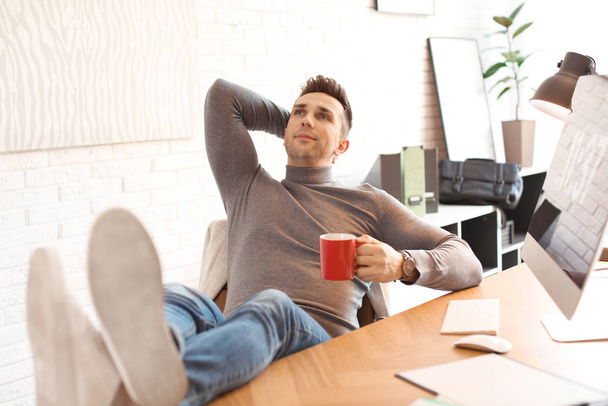 Young man with cup of drink relaxing at table in office during b - Foto, Imagen