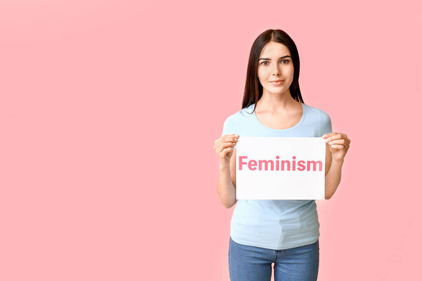 Mujer joven sosteniendo papel con texto FEMINISMO sobre fondo de color
 - Foto, Imagen