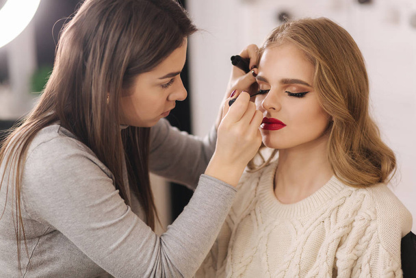 Makeup artist work in beauty studio. Woman applying by professional make up master. Beautiful make up artist make a makeup for blond hair model - 写真・画像